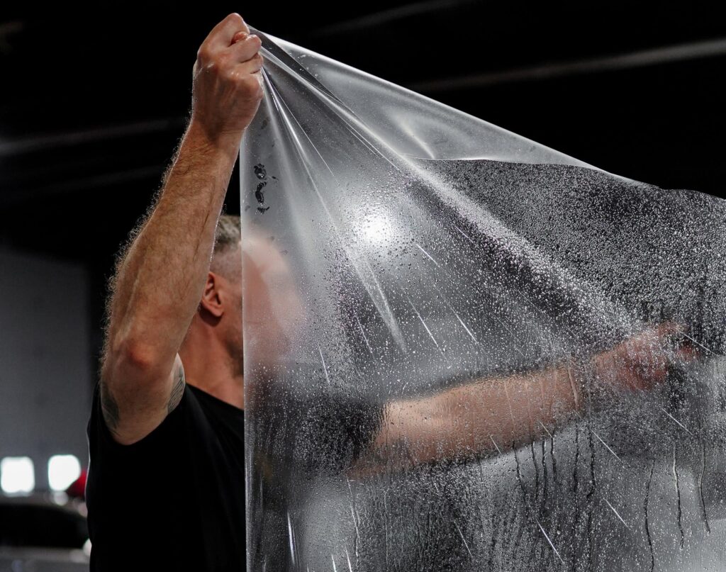 Close-up of a person lifting a clear plastic sheet, with water droplets clinging to its surface against a dark background.